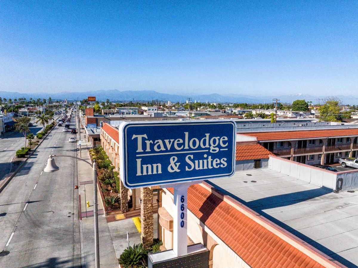 An aerial view of a Travelodge Inn & Suites with a large blue sign in the foreground, located alongside a main road with mountains in the distance.