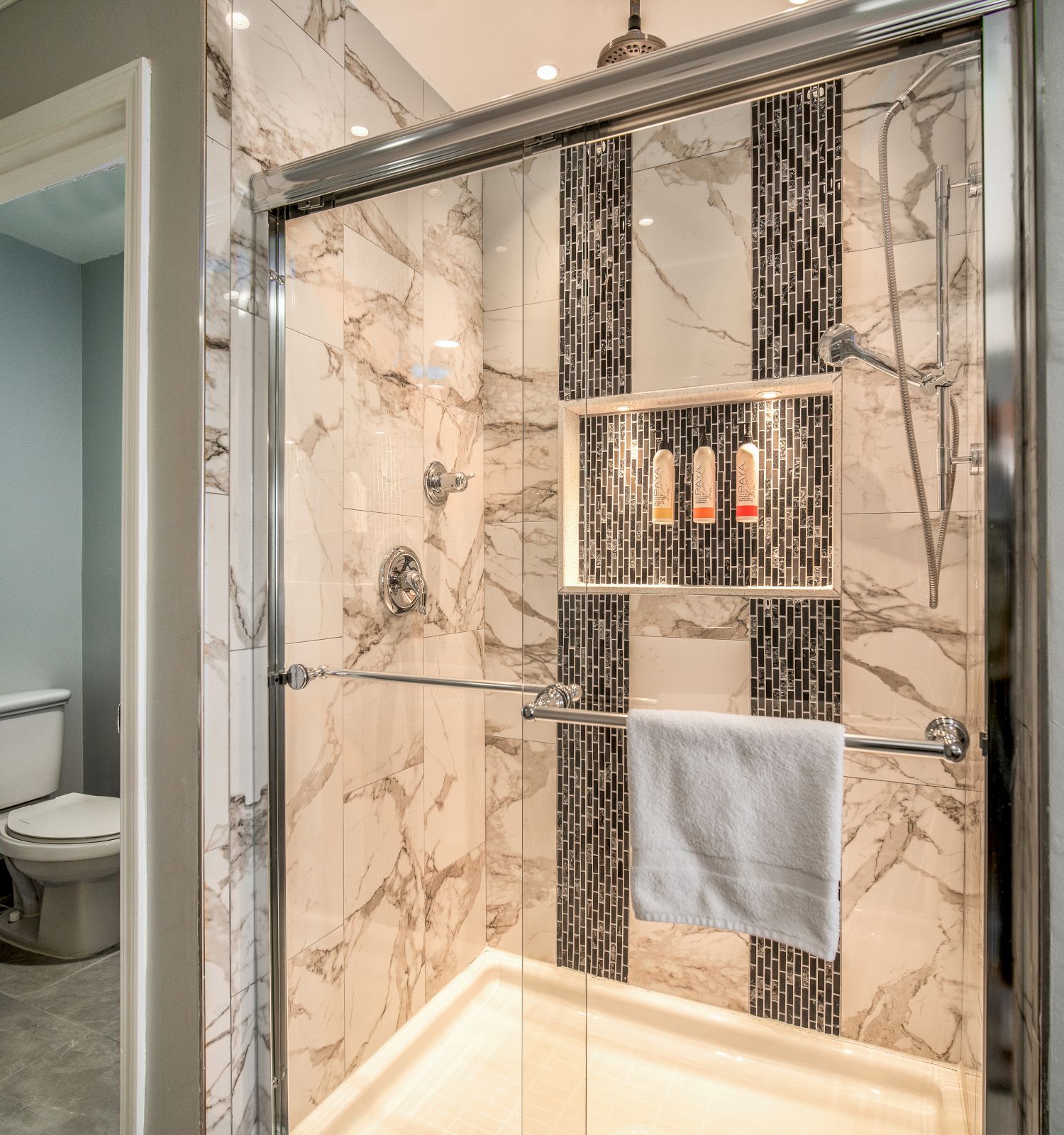 The image shows a modern bathroom with a glass-enclosed shower featuring marble and mosaic tiles, a towel hanging on the door, and a view of the toilet.