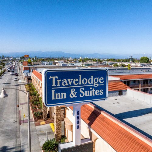 The image shows a Travelodge Inn & Suites sign in front of a hotel, with a street and buildings stretching into the distance on a clear day.