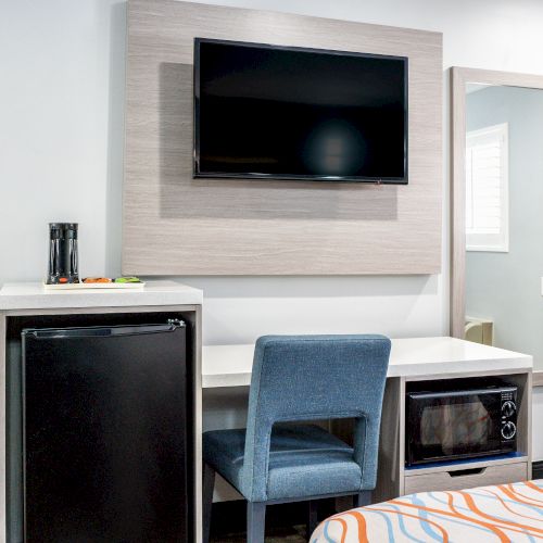 A hotel room setup with a wall-mounted TV, a desk, a blue chair, a mini-fridge, a microwave, and a mirror next to a small window.