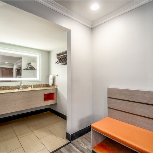 The image shows a modern bathroom with a sink, mirror, towel rack, hairdryer, and an orange bench with a wooden backrest under a lighted ceiling.