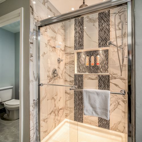 The image shows a modern bathroom with a glass-enclosed shower, marble tiles, a built-in shelf with toiletries, and a towel hanging on the door. A toilet is visible.