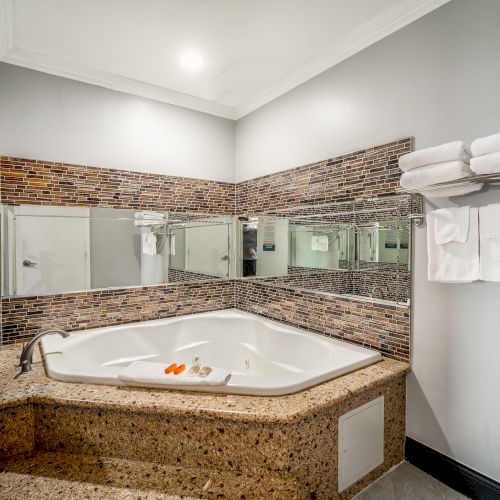 This image shows a bathroom with a large corner bathtub, mirrors, a tiled countertop, and a towel rack with folded towels on the wall.