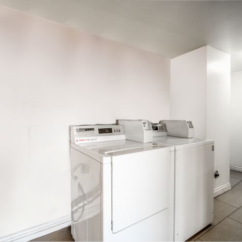 The image shows a clean laundry room with two white washing machines against a white wall and gray tiled floor.