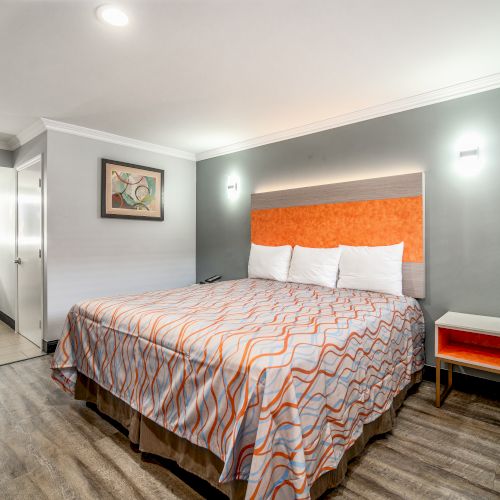 A modern hotel room features a king-sized bed with an orange and gray theme, nightstand, chair, wall art, and a view into the bathroom.