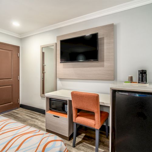 The image shows a hotel room with a brown door, a wall-mounted TV, a desk with an orange chair, a mirror, and a mini fridge.