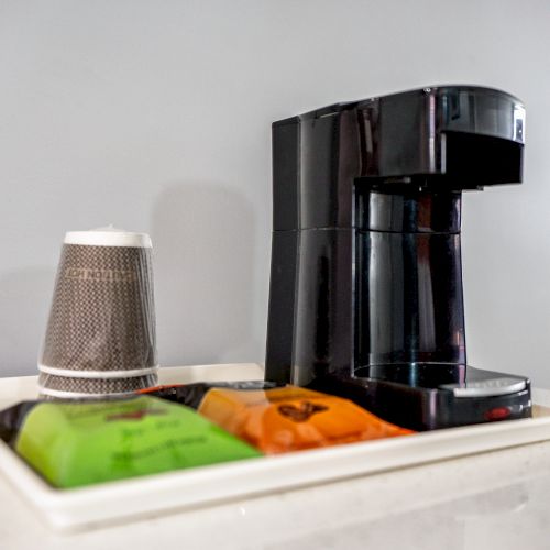 The image shows a coffee maker on a tray with disposable coffee cups and coffee packets on a counter, providing a convenient setup for making coffee.
