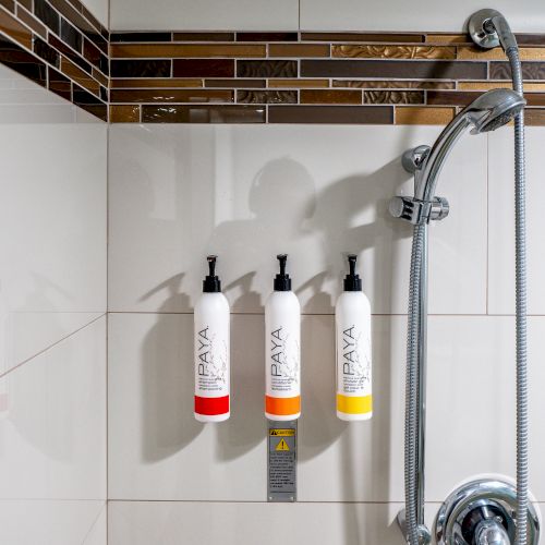 A shower setup with three wall-mounted bottles of bathroom products and a handheld showerhead, set against tiled walls.