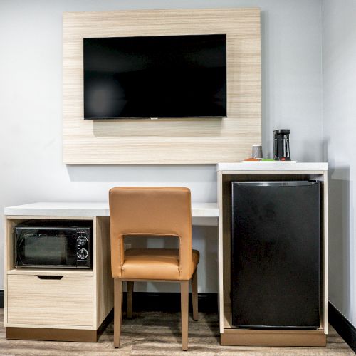 A small room setup featuring a wall-mounted TV, a desk with a chair, a microwave, a mini-fridge, and a coffee maker on a countertop.