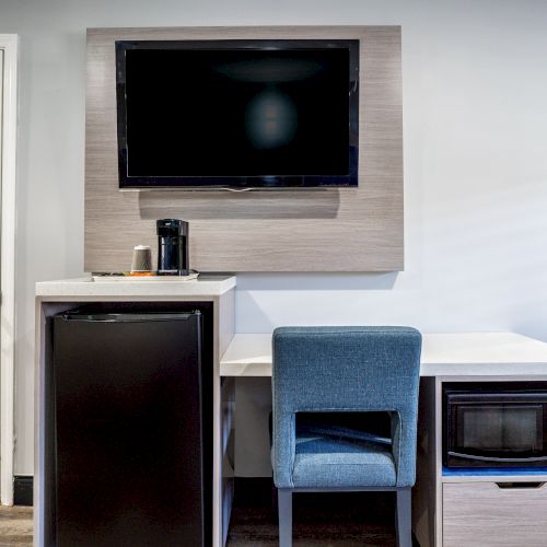 A modern room with a wall-mounted TV, a desk, a blue chair, a mini-fridge, a coffee maker, a microwave, a mirror, and a closed door is shown.