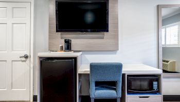 The image shows a modern hotel room setup with a wall-mounted TV, mini-fridge, coffee maker, blue chair, small desk, and mirror.