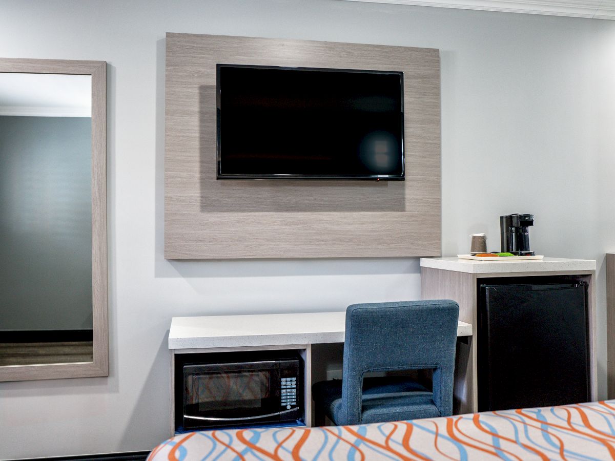 The image shows a hotel room with a TV, desk, chair, mirror, mini-fridge, microwave, and a coffee maker on a counter.