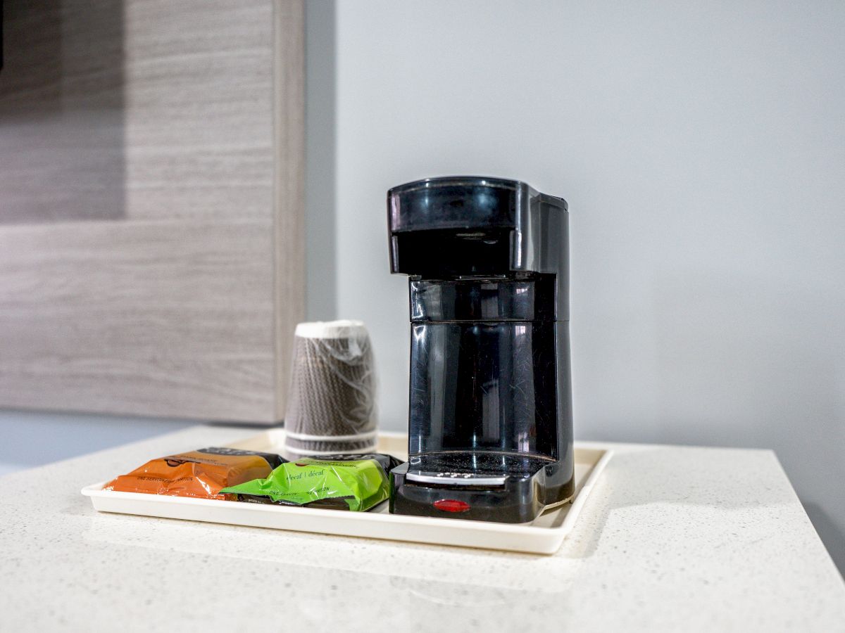 A single-cup coffee maker on a countertop, with a tray holding plastic-wrapped disposable cups and coffee packets in various colors.